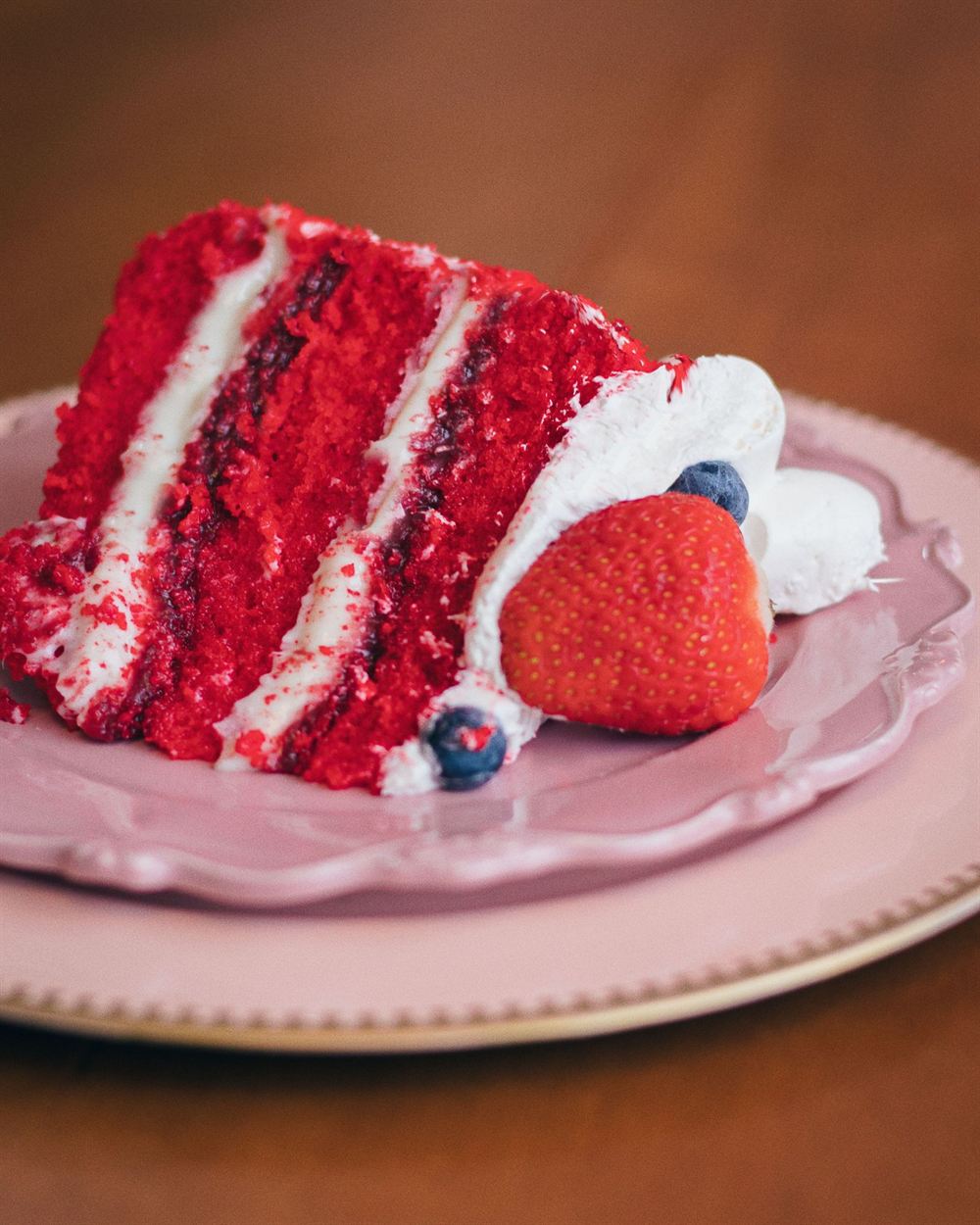 Fatia de bolo red velvet com recheio de cream cheese, morango e mirtilo num prato rosa