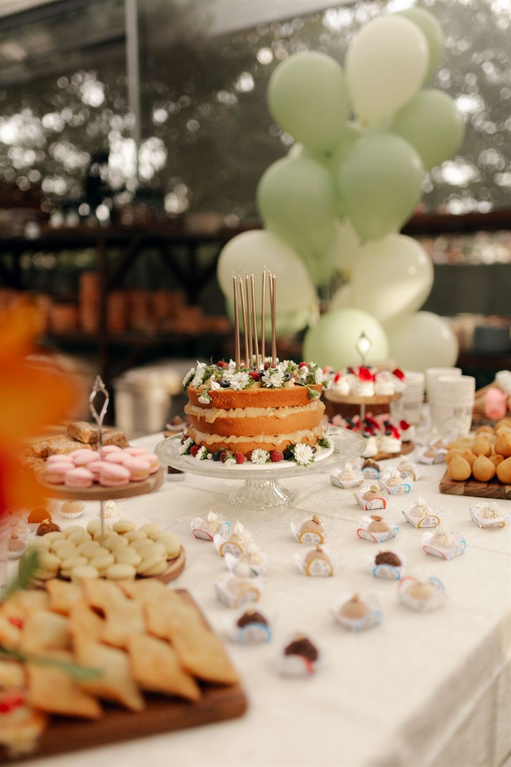 Mesa de festa com naked cake de velas altas, macarons, docinhos em forminhas e balões verdes ao fundo