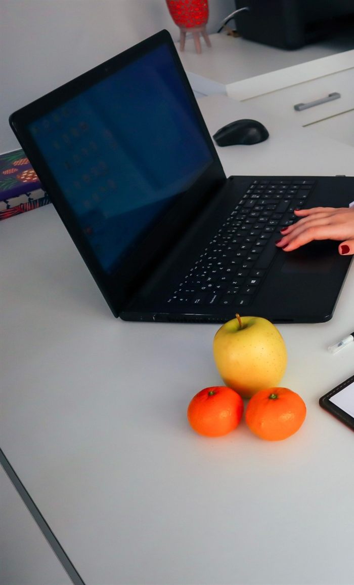 Mesa do consultório com notebook, uma maçã e duas tangerinas, e a mão da nutricionista no teclado
