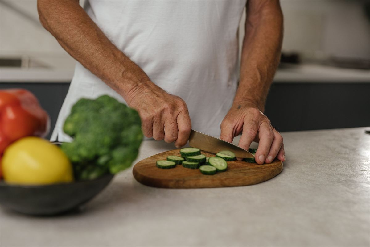 Mãos cortando pepino numa tábua, com brócolis e pimentões ao lado