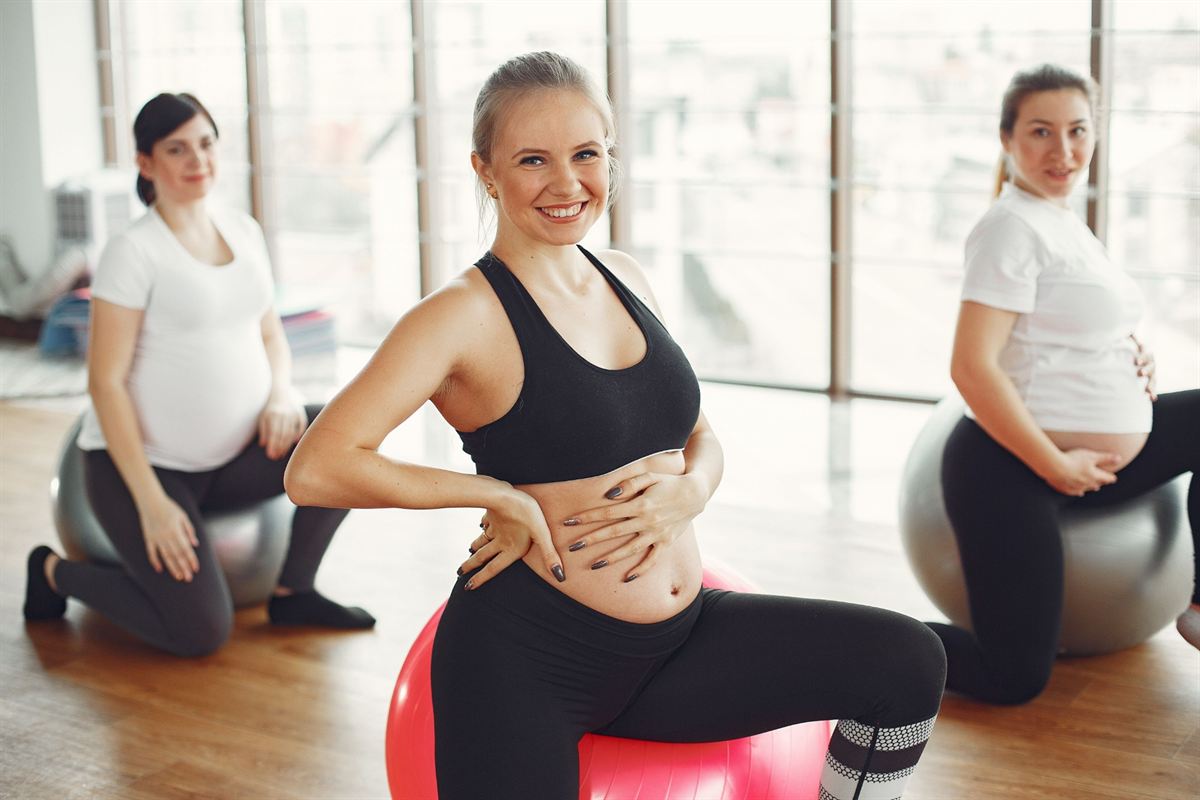 Três gestantes sentadas em bolas de pilates numa sala com janelas grandes